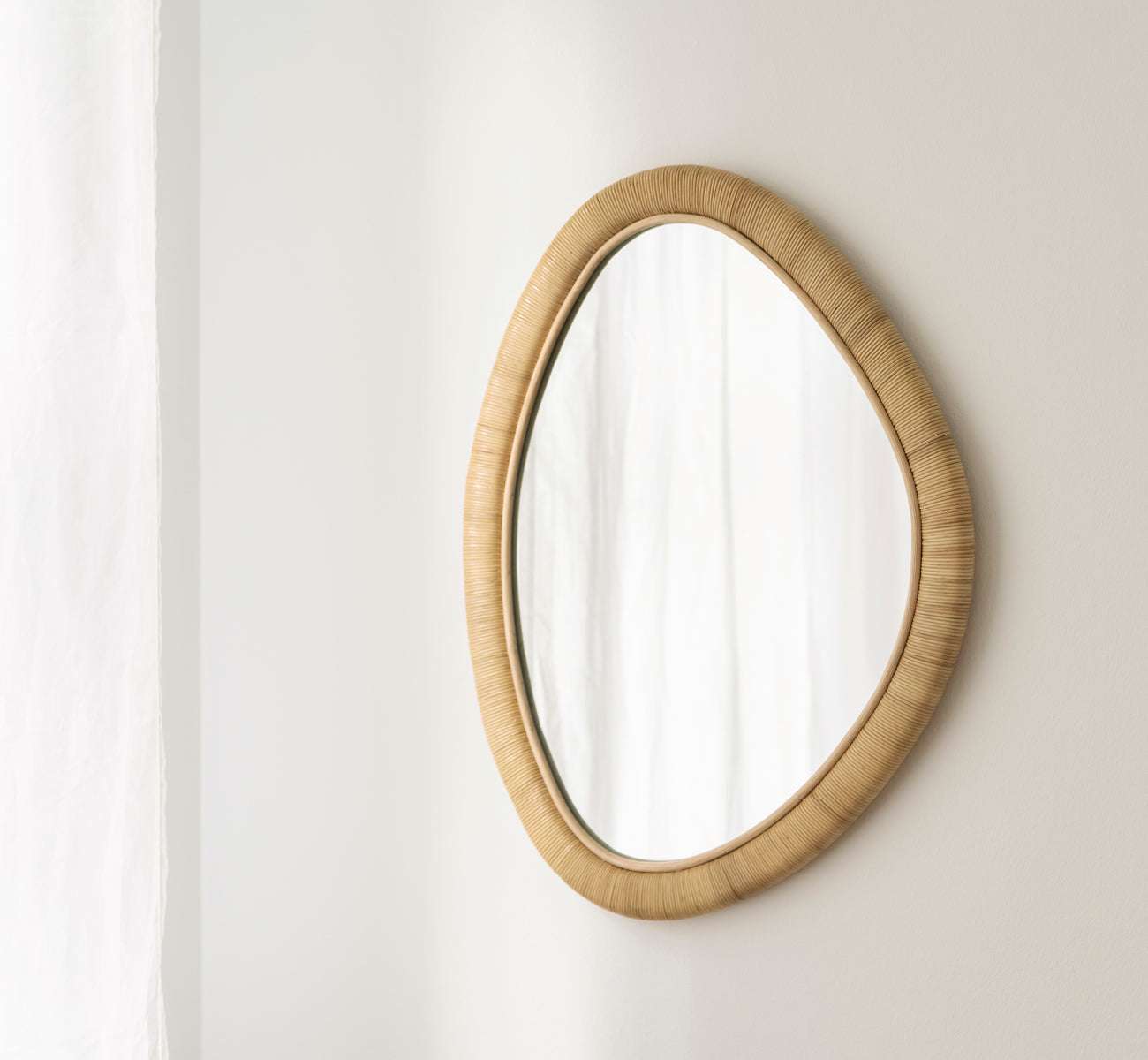 Esta es una imagen de estilo de vida de Malou espejo en natural rattan. Forma ovalada orgánica montada en una suave pared blanca con cortina.