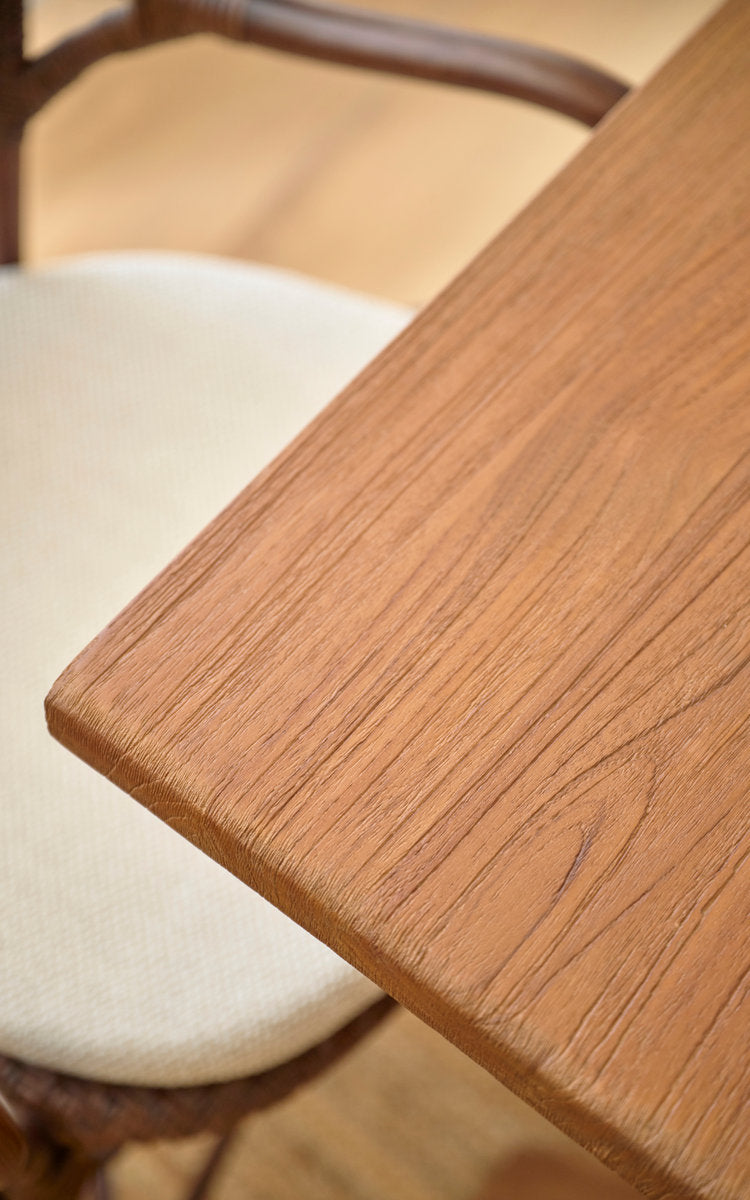 Esta es una imagen de detalle de Hugo mesa de comedor de teca 140x90 cm en teak indoor. Borde liso de la tapa de teca junto a una silla de ratán.