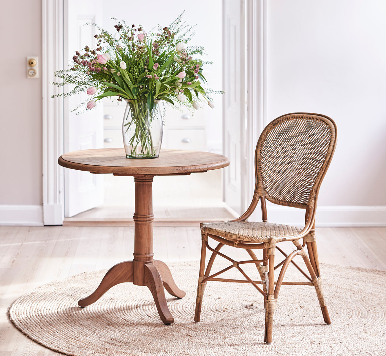 Esta es una imagen de estilo de vida de Rossini Dining Chair i antique rattan. Colocada alrededor de una mesa redonda de teca sobre una alfombra de yute en un loft luminoso.