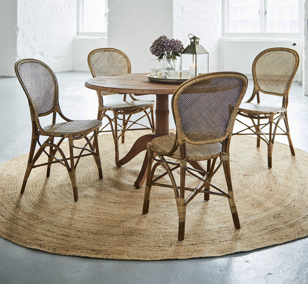 Esta es una imagen de estilo de vida de Rossini Dining Chair i antique rattan. Cuatro sillas alrededor de una mesa redonda de teca sobre una alfombra de yute en un loft blanco.
