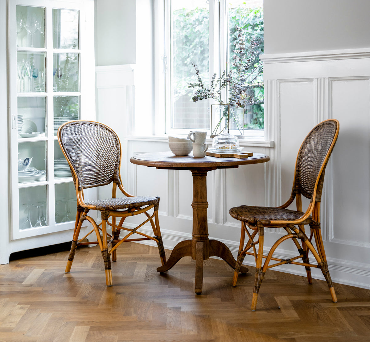 Esta es una imagen de estilo de vida de Rossini Dining Chair i cherry rattan. Dos sillas junto a una mesa pequeña en un patio con suelo de baldosas ajedrezadas.