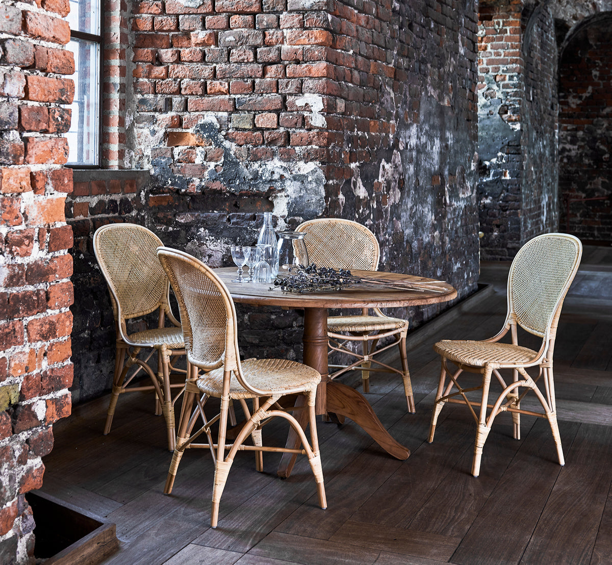 Esta es una imagen de estilo de vida de Rossini silla de comedor en natural rattan. Colocada junto a una mesa bistró de vidrio sobre una alfombra de yute en un pasillo luminoso.