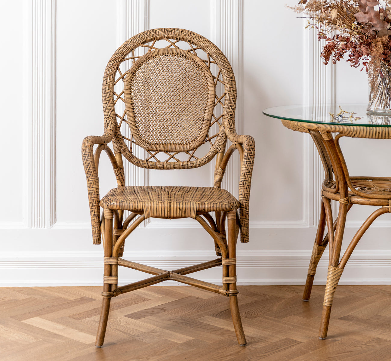 Esta es una imagen de estilo de vida de Romantica silla de comedor en antique rattan. Colocada junto a una mesa de cristal y ratán con flores secas en una sala con paneles.