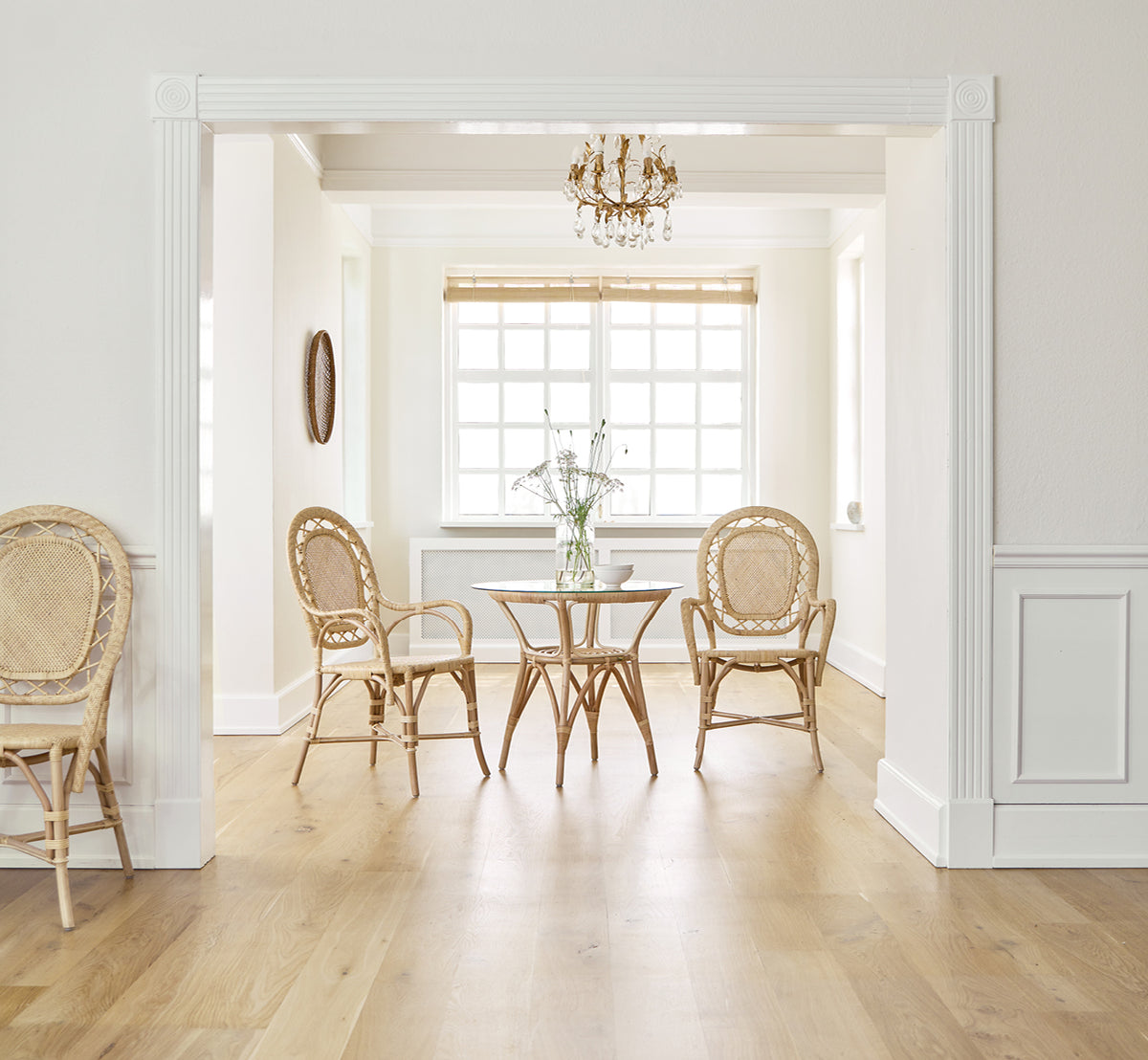Esta es una imagen de estilo de vida de Romantica silla de comedor en natural rattan. Alrededor de una mesa redonda en una sala elegante con araña y paneles blancos.