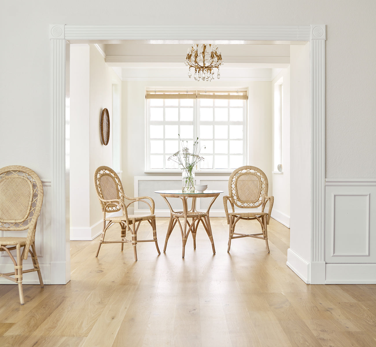 Esta es una imagen de estilo de vida de Romantica silla de comedor en natural rattan. Alrededor de una mesa redonda en una sala elegante con araña y paneles blancos.