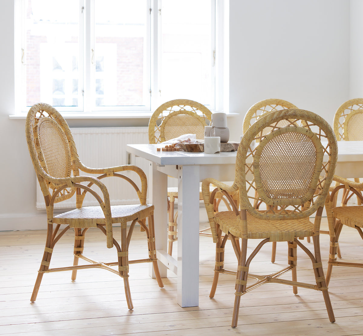 Esta es una imagen de estilo de vida de Romantica silla de comedor en natural rattan. Reunidas alrededor de una mesa de comedor blanca en una luminosa sala escandinava.