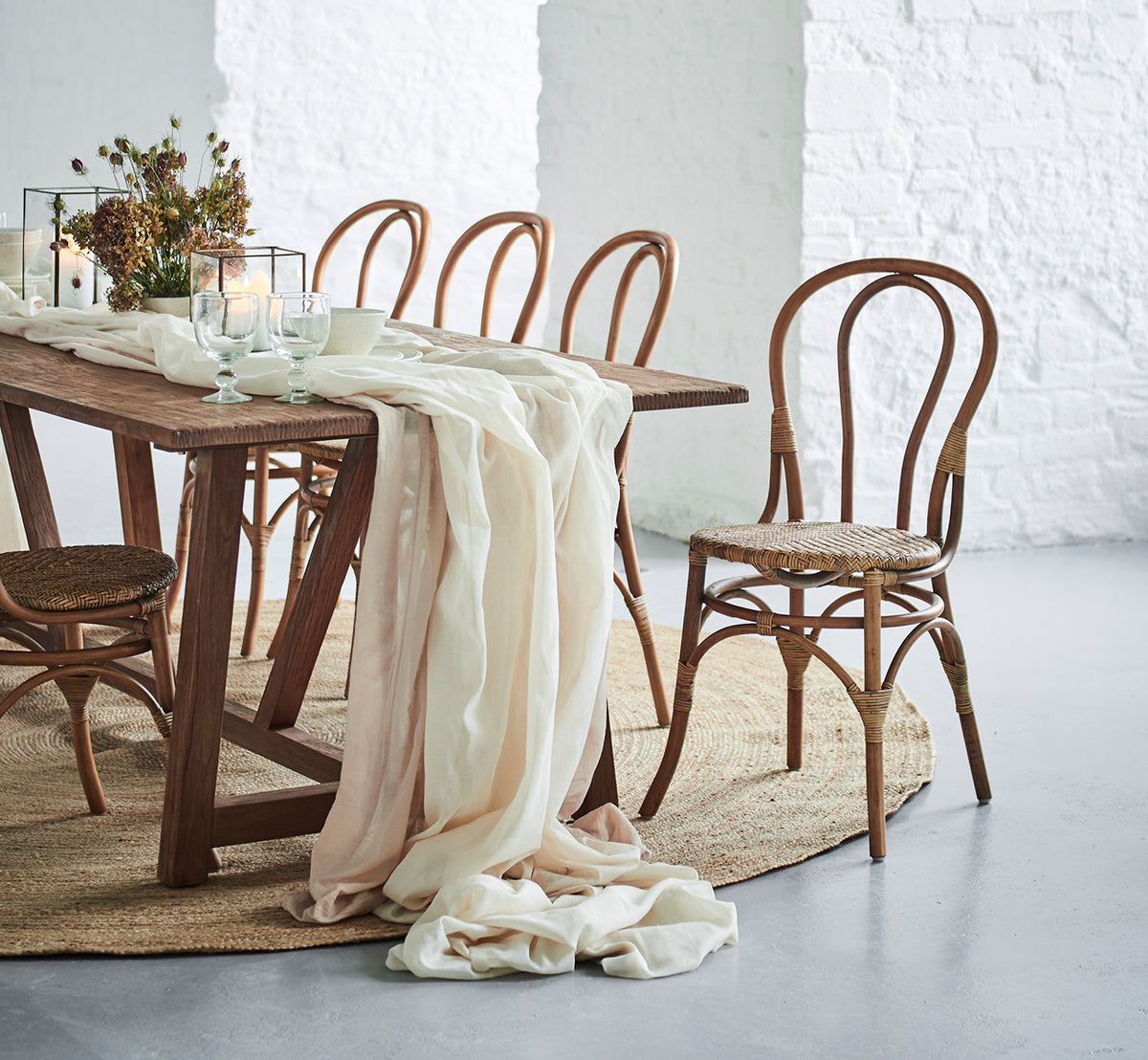 Esta es una imagen de estilo de vida de Lulu silla de comedor en antique rattan. En una mesa rústica con lino drapeado y velas contra una pared de ladrillo blanco.