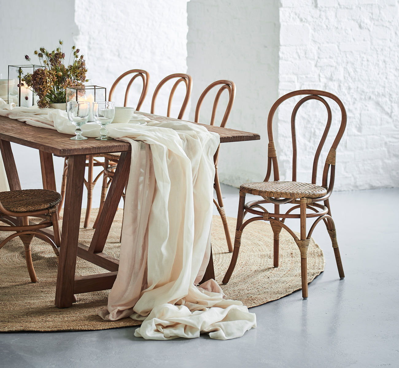 Esta es una imagen de estilo de vida de Lulu silla de comedor en antique rattan. En una mesa rústica con lino drapeado y velas contra una pared de ladrillo blanco.