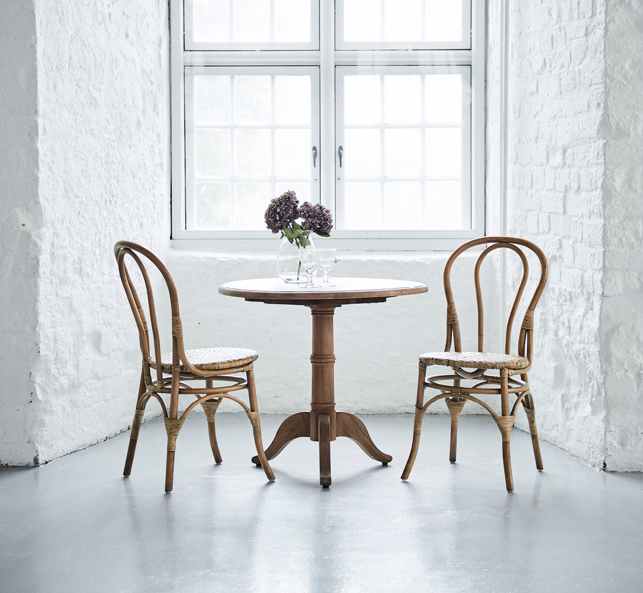 Esta es una imagen de estilo de vida de Lulu silla de comedor en antique rattan. Dos sillas flanquean una mesa redonda con flores en un interior de ladrillo blanco luminoso.