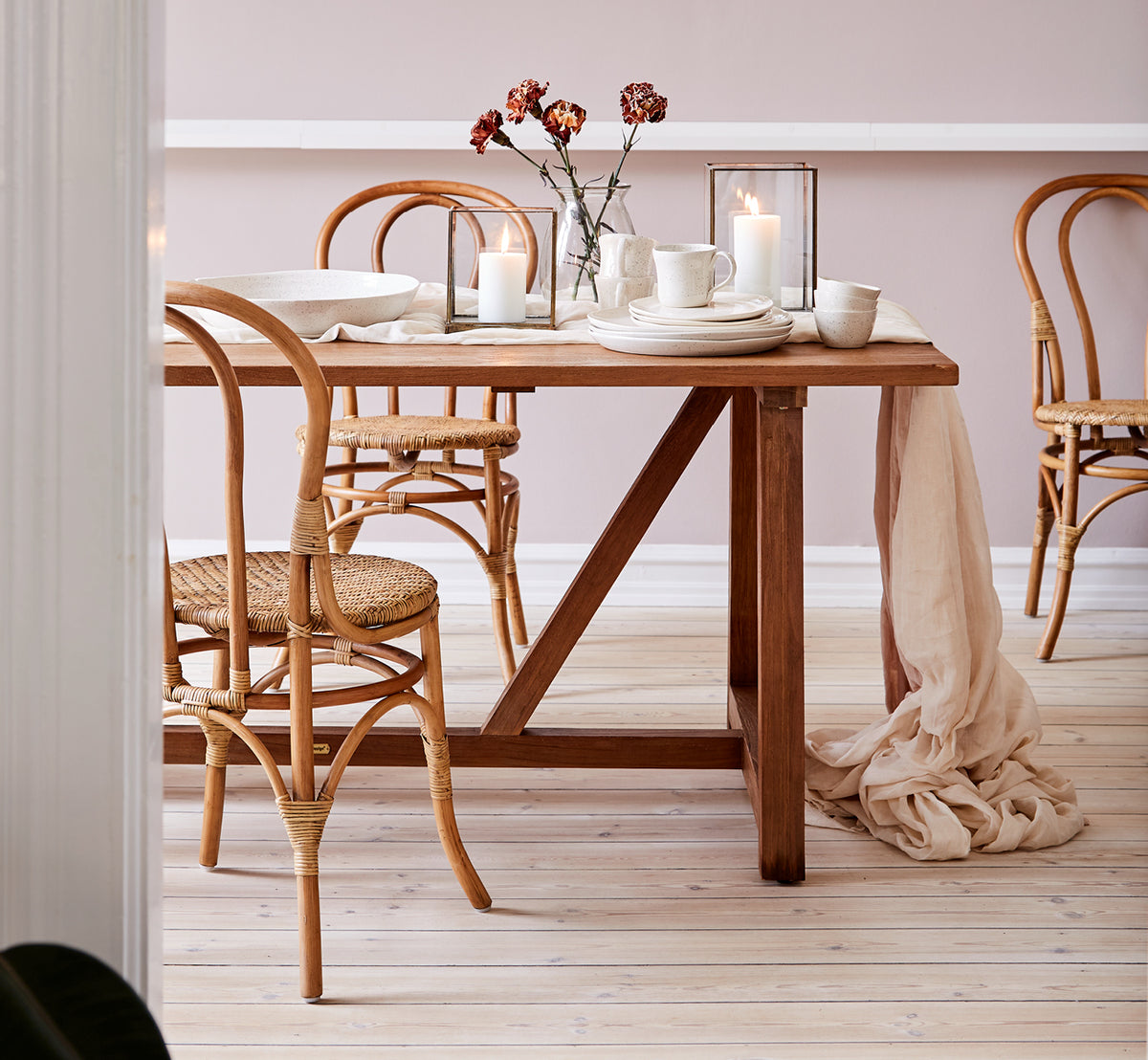 Esta es una imagen de estilo de vida de Lulu silla de comedor en antique rattan. Estilizada junto a una mesa de comedor de teca con velas y lino en un interior cálido.