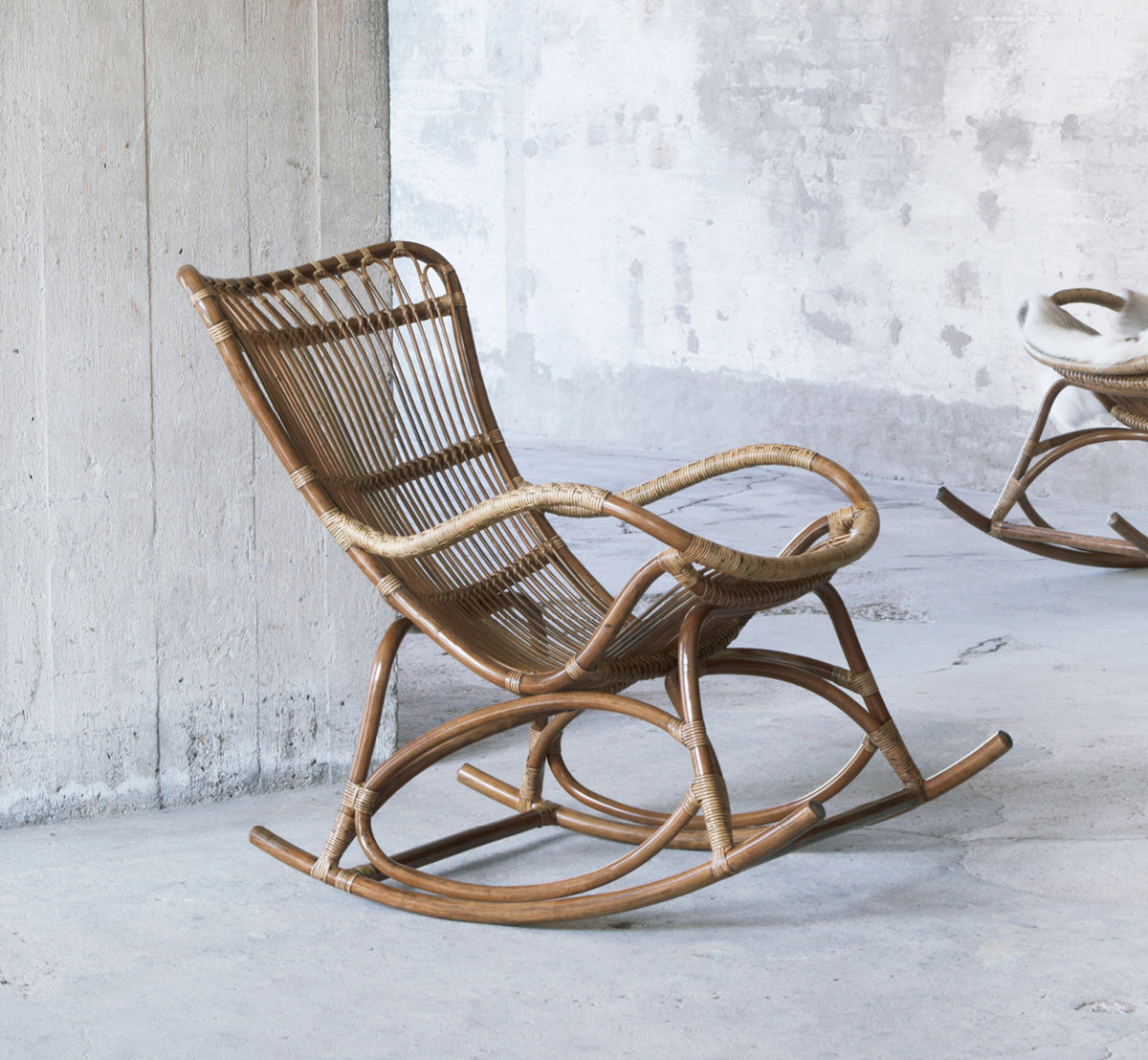 Esta es una imagen de estilo de vida de Monet Rocking Chair i antique rattan. En una sala rústica de hormigón con otra mecedora al fondo.