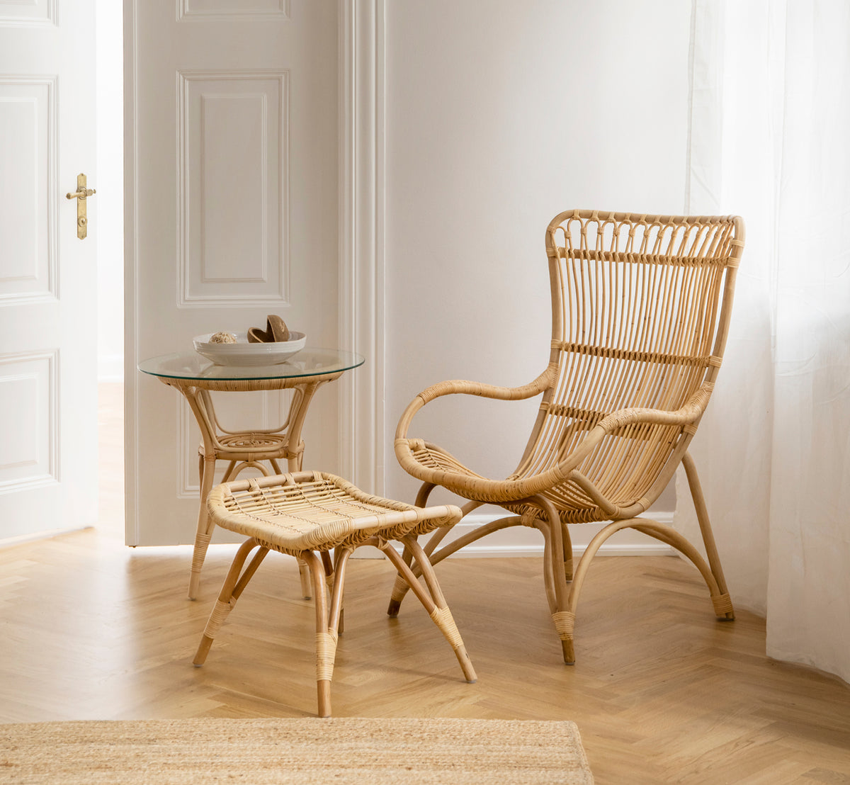 Esta es una imagen de estilo de vida de Monet Lounge Chair i natural rattan. Con reposapiés y mesa auxiliar junto a una puerta sobre suelo en espiga.