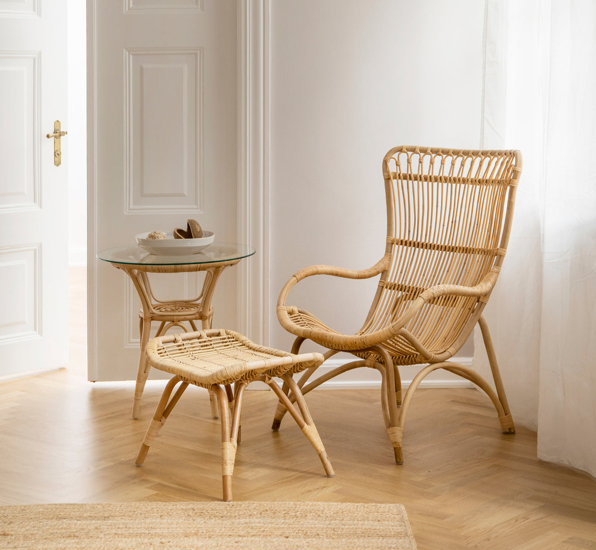 Esta es una imagen de estilo de vida de Monet reposapiés en natural rattan. Combinada con sillón de ratán de respaldo alto y mesa auxiliar de vidrio en una entrada luminosa.