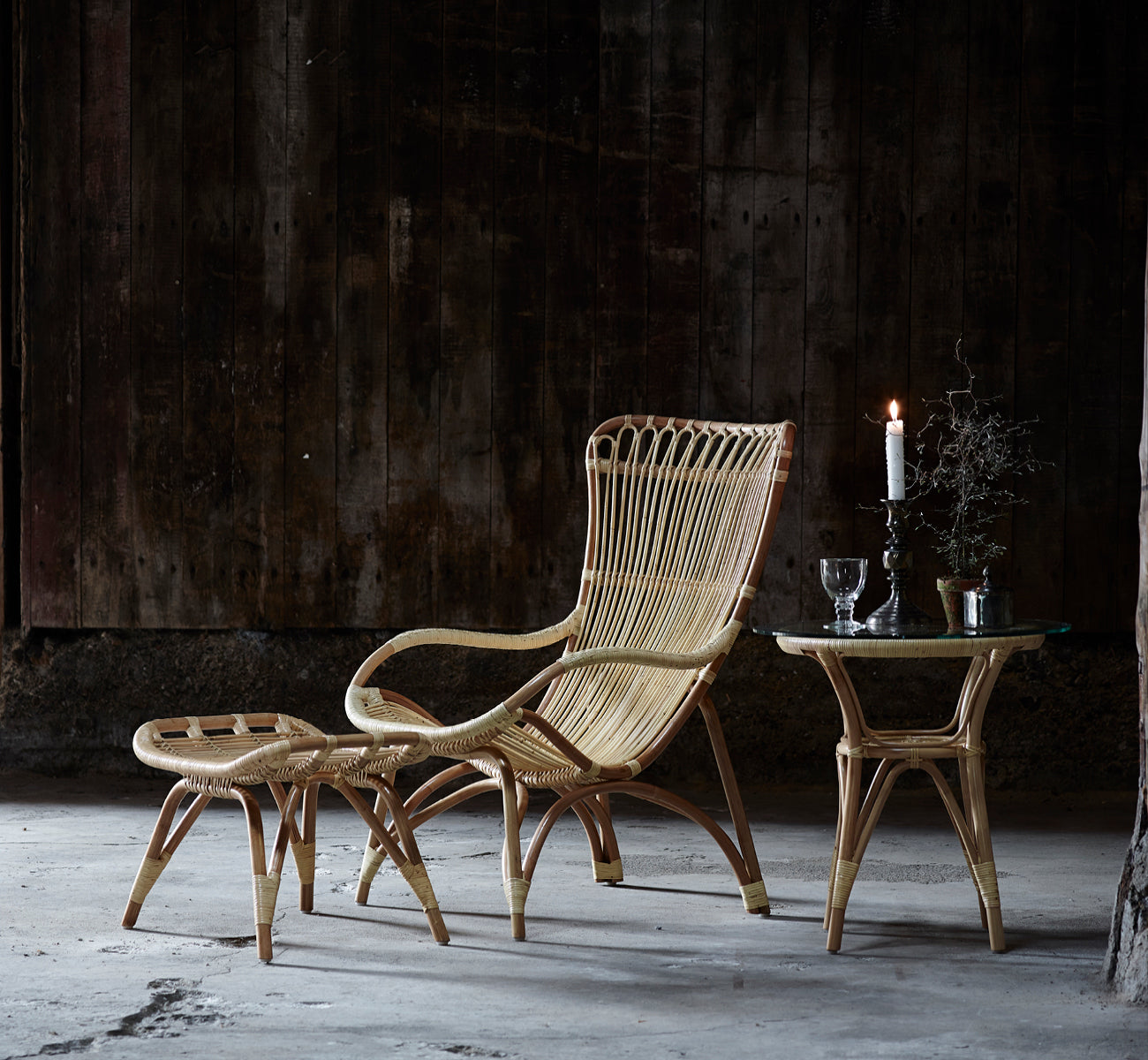 Esta es una imagen de estilo de vida de Monet Footstool i natural rattan. Con sillón lounge en un ambiente rústico de granero oscuro con cristalería.
