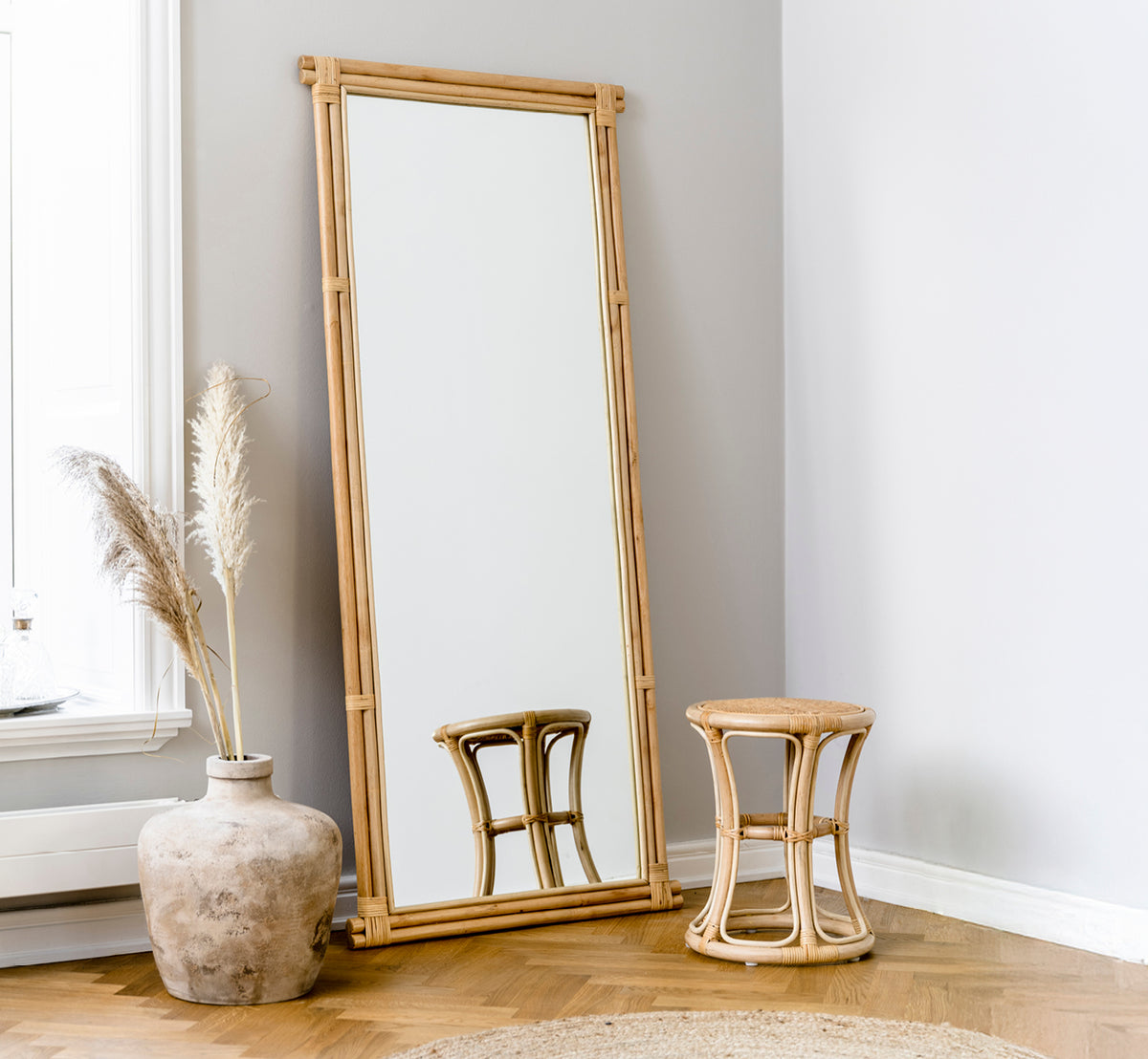 Esta es una imagen de estilo de vida de Rasmus Mirror 172 cm en natural rattan. Apoyado contra una pared gris junto a un taburete y hierba de las pampas seca.