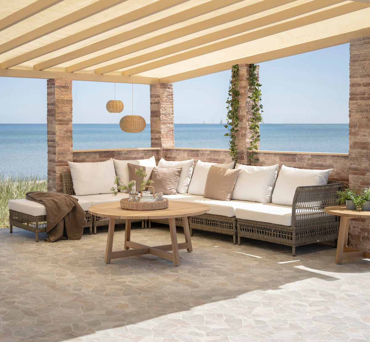Esta es una imagen de estilo de vida de Mona sofá modular exterior-Center Module en antique grey artfibre aluminium. Bajo una pérgola junto al mar.