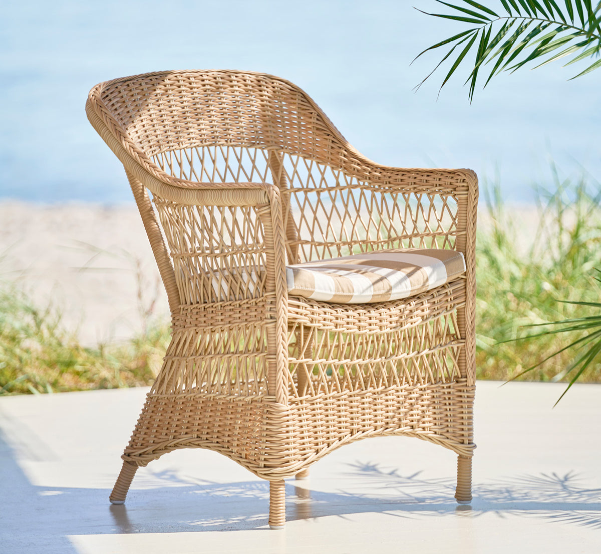 Esta es una imagen de estilo de vida de Charlot silla en natural ArtFibre Aluminium, en una terraza costera soleada con vistas al mar y una pérgola.