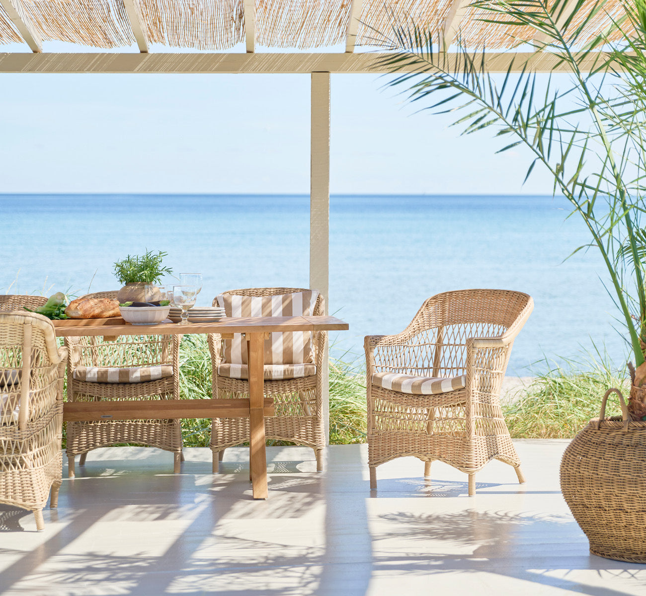 Esta es una imagen de estilo de vida de Charlot silla en natural ArtFibre Aluminium, en una terraza costera soleada con vistas al mar y una pérgola.