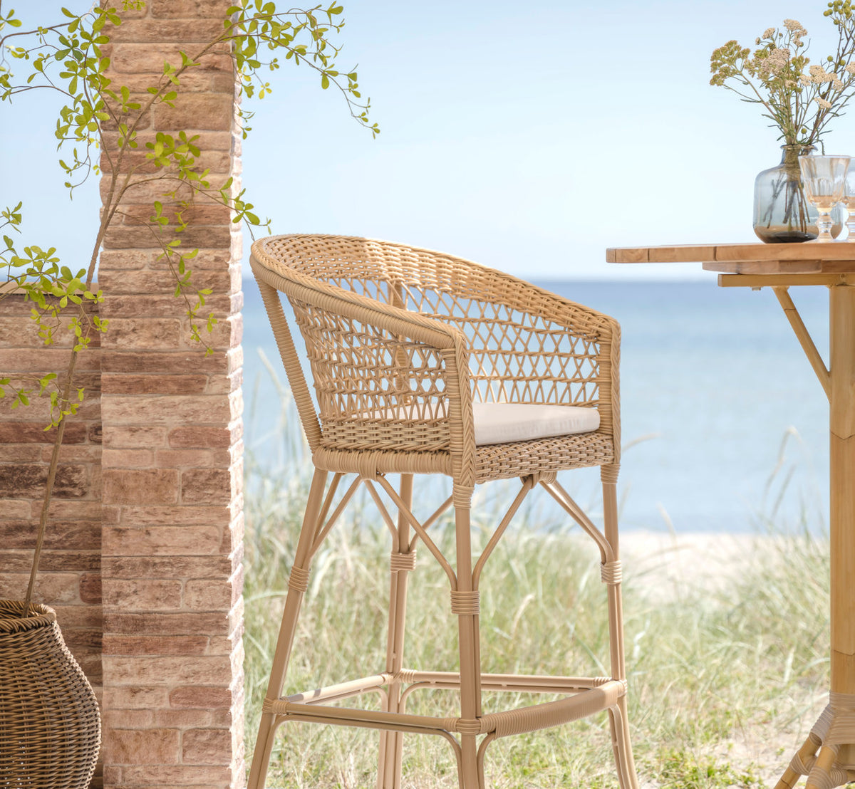 Esta es una imagen de estilo de vida de Emma taburete de bar exterior en natural artfibre aluminium. Junto a un pilar de ladrillo con hierba de duna y vista al mar.