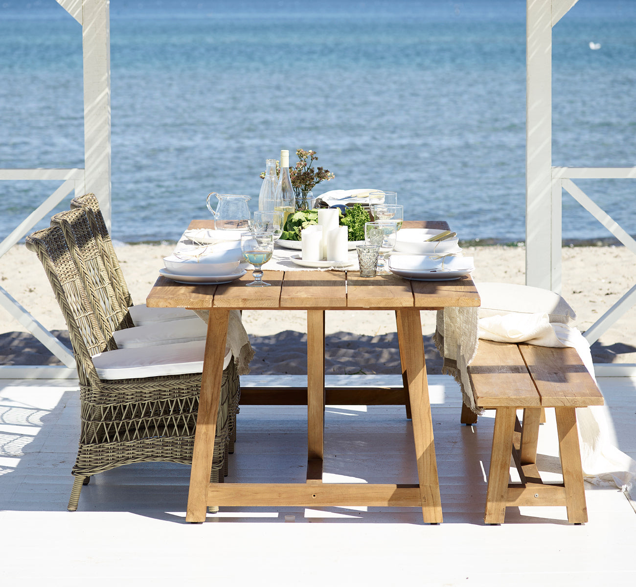 Esta es una imagen de estilo de vida de Marie Exterior Side Chair i antique grey artfibre aluminium. Junto a una mesa de teca en un pabellón junto al mar.