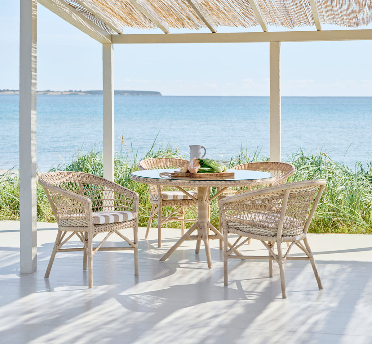 Esta es una imagen de estilo de vida de Emma silla de comedor exterior en natural ArtFibre Aluminium, en una terraza costera luminosa con techo de.