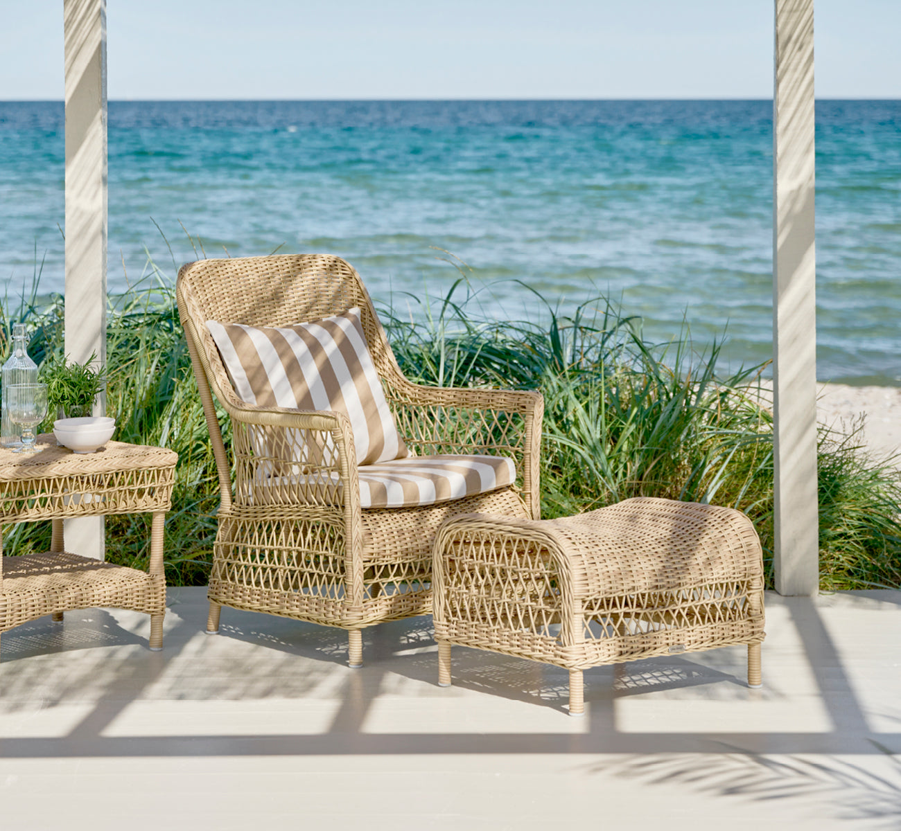 Esta es una imagen de estilo de vida de Dawn reposapiés en natural ArtFibre Aluminium, en una terraza costera con vistas al mar, gramíneas de playa y.