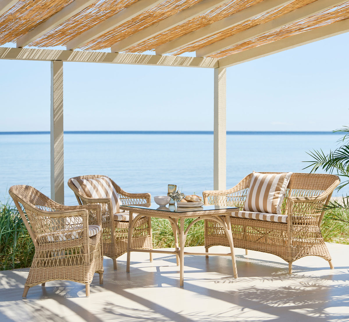 Esta es una imagen de estilo de vida de Charlot sofá de 2 plazas en natural ArtFibre Aluminium, en una terraza costera con techo de caña, vistas al.