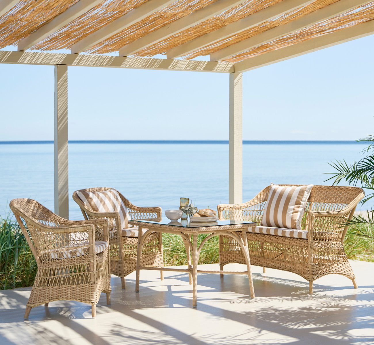 Esta es una imagen de estilo de vida de Charlot sofá de 2 plazas en natural ArtFibre Aluminium, en una terraza costera con techo de caña, vistas al.