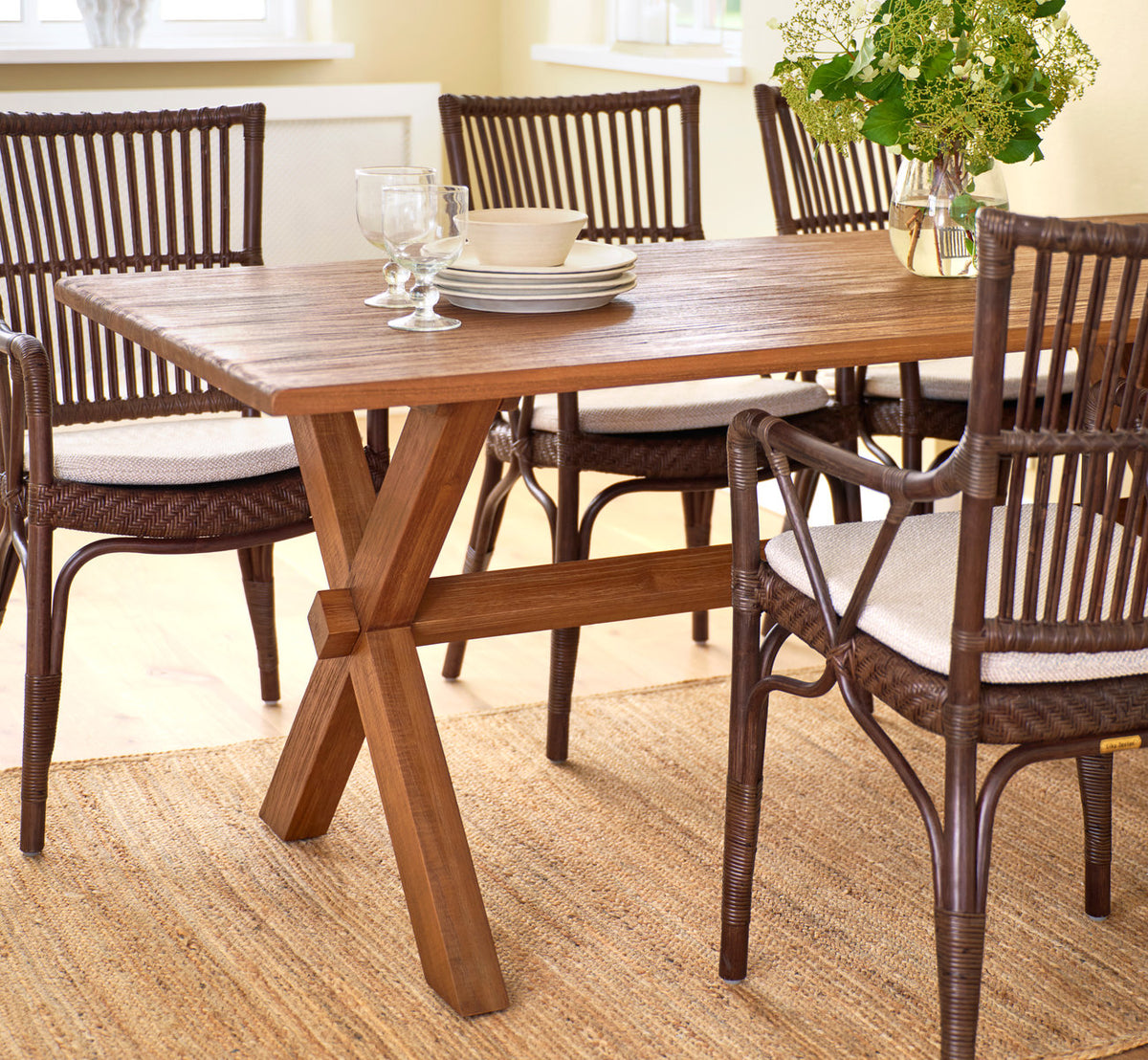 Esta es una imagen de estilo de vida de Hugo mesa de comedor de teca 180x90 cm en teak indoor. Con sillas de comedor de ratán sobre alfombra de yute.