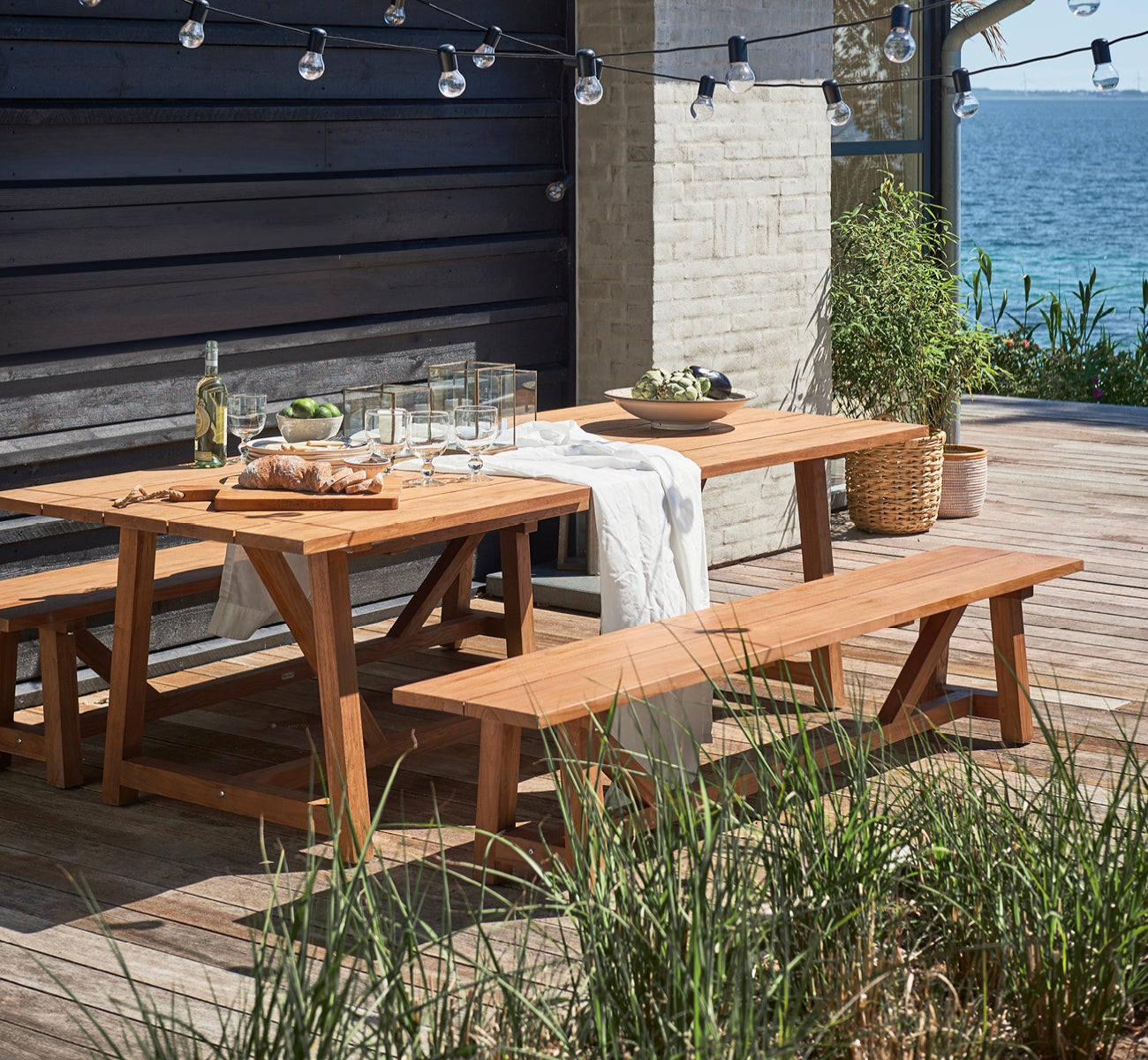 Esta es una imagen de estilo de vida de George mesa exterior 240 en outdoor Teak, en un patio de jardín exterior con fachada blanca y vegetación.