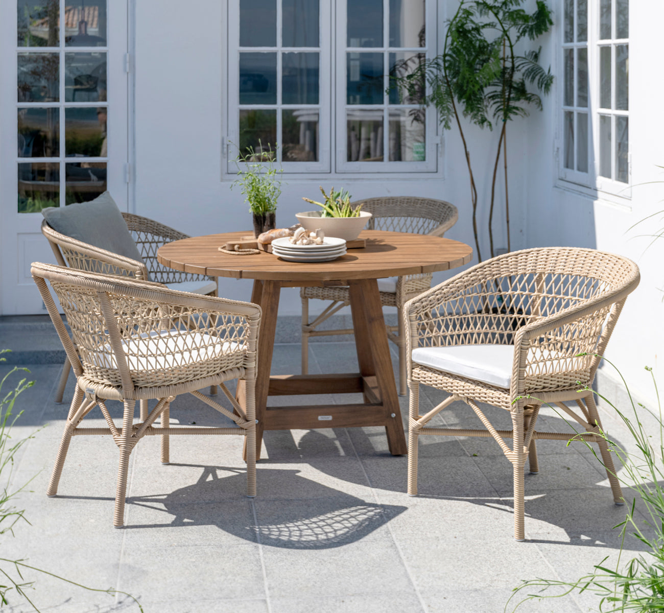 Esta es una imagen de estilo de vida de George mesa exterior Ø120 en outdoor Teak, en un patio de jardín exterior con fachada blanca y vegetación.