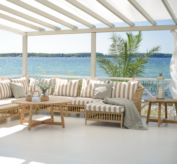 Esta es una imagen de estilo de vida de Victor Teak Lounge Table Ø120 cm en teak outdoor. Colocada entre sofás exteriores con cojines a rayas con vistas al mar.