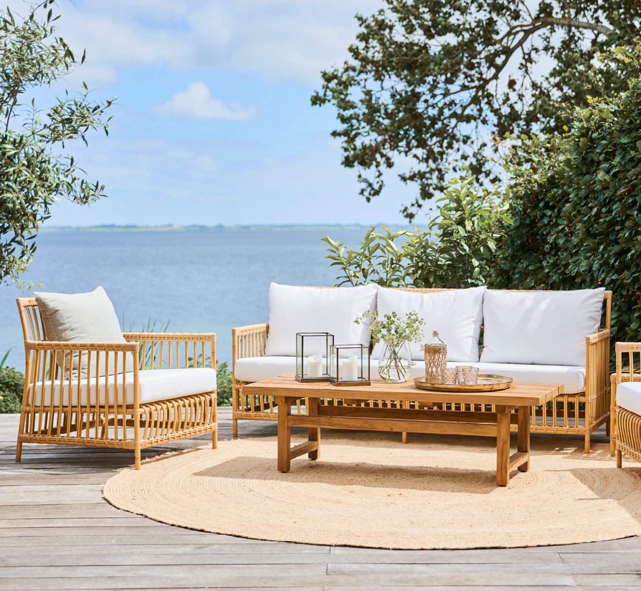 Esta es una imagen de estilo de vida de Julian Teak Lounge Table 140x55 cm en teak outdoor. En una terraza con muebles lounge, una alfombra de yute y vistas al mar.