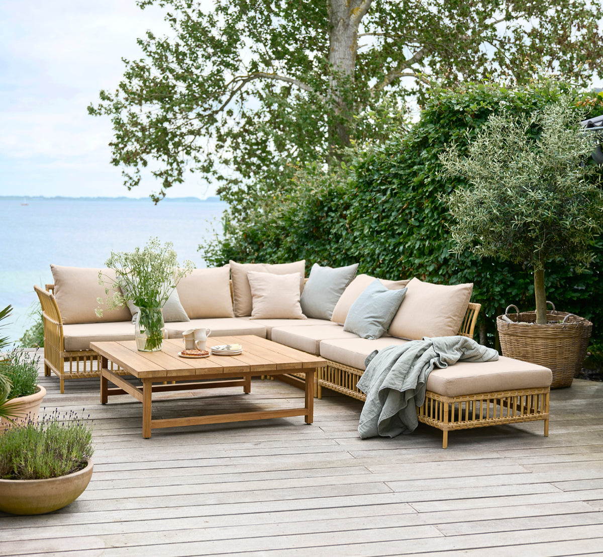 Esta es una imagen de estilo de vida de Julian Teak Lounge Table 120x120 cm en teak outdoor. En una terraza de jardín junto al mar con asientos lounge y exuberante vegetación.