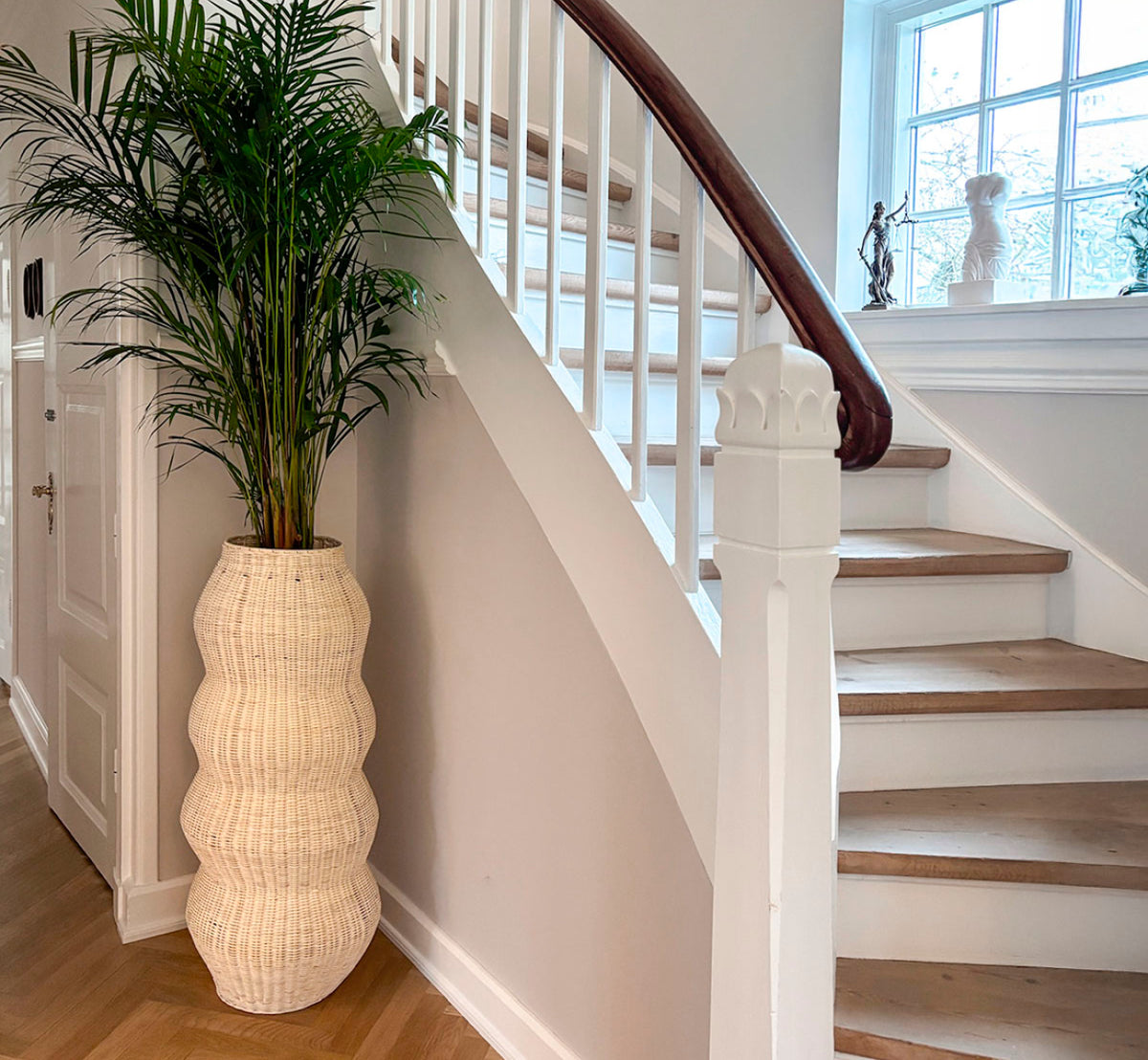 Esta es una imagen de estilo de vida de Unda Basket H102 cm en natural core weave iron frame. Utilizada como macetero alto junto a una escalera curva en un vestíbulo luminoso.