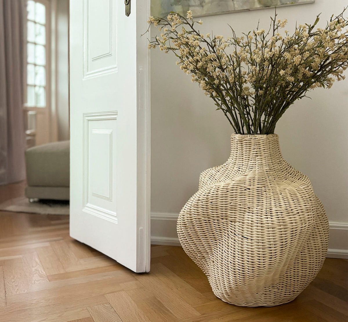 Esta es una imagen de estilo de vida de Solaris Basket Ø46 cm en natural core weave iron frame. Estilizada con flores secas junto a una puerta blanca sobre un suelo de parqué.