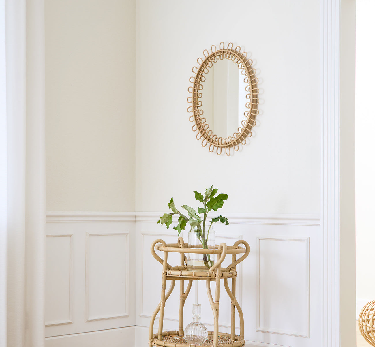 Esta es una imagen de estilo de vida de Luella espejo en polished natural rattan. Montado en una pared con paneles blancos sobre un carrito bar de ratán con plantas.