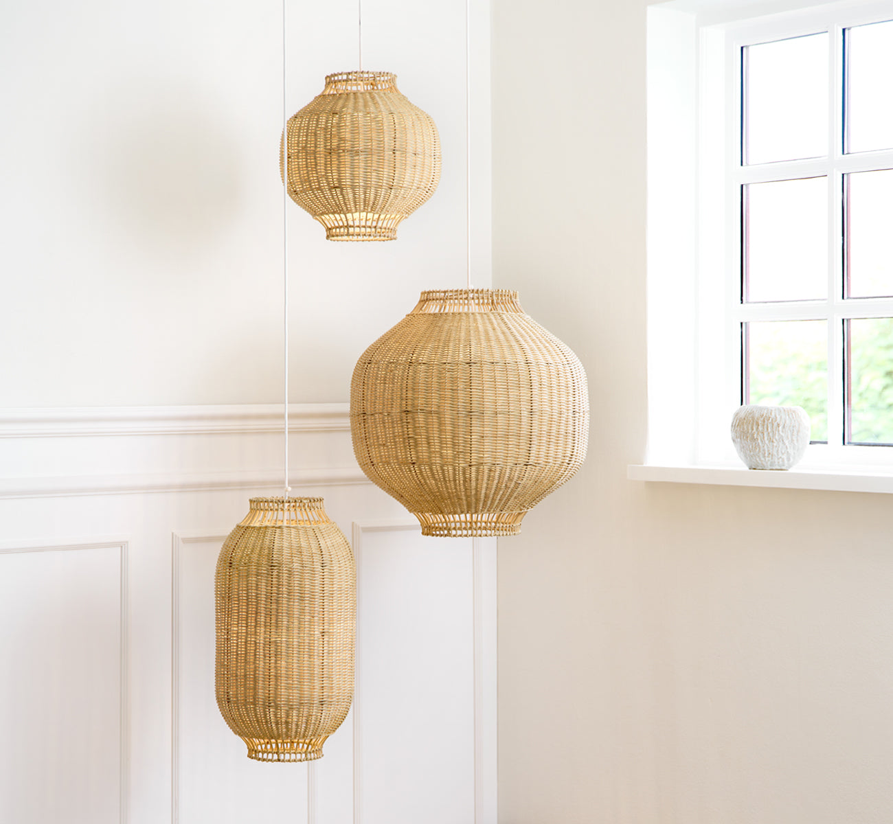 Esta es una imagen de estilo de vida de Kou pantalla de lámpara en natural core weave rattan frame. Tres tamaños cuelgan juntos en una luminosa sala con paneles blancos.
