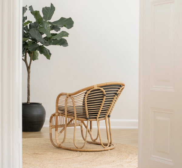 Esta es una imagen de estilo de vida de Swing sillón lounge en polished natural rattan. De pie sola junto a una planta frondosa en un suelo de parqué en espiga.