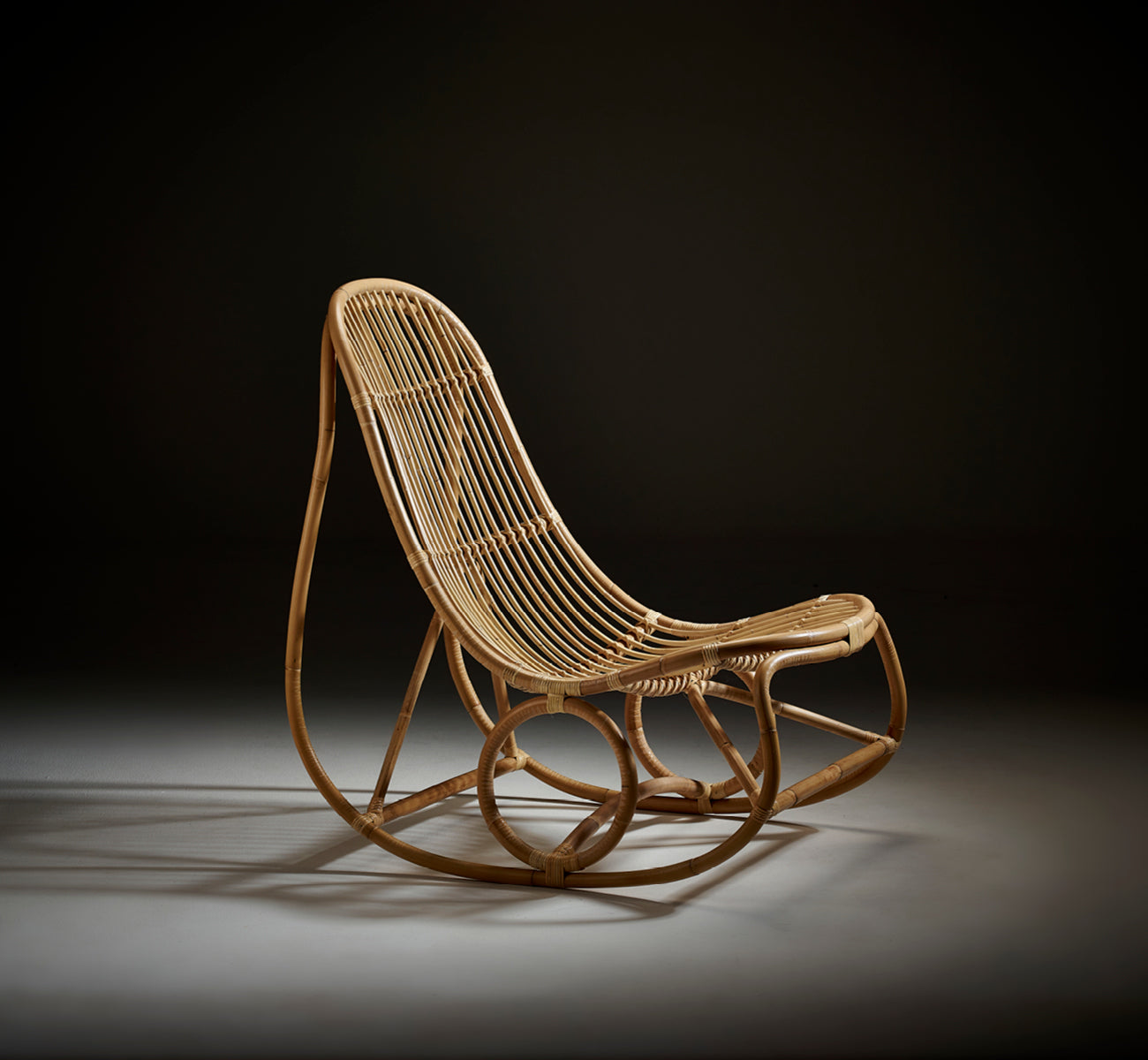 Esta es una imagen de estilo de vida de Nanny Rocking Chair i polished natural rattan. Foto de estudio sobre fondo oscuro que destaca sus curvas.