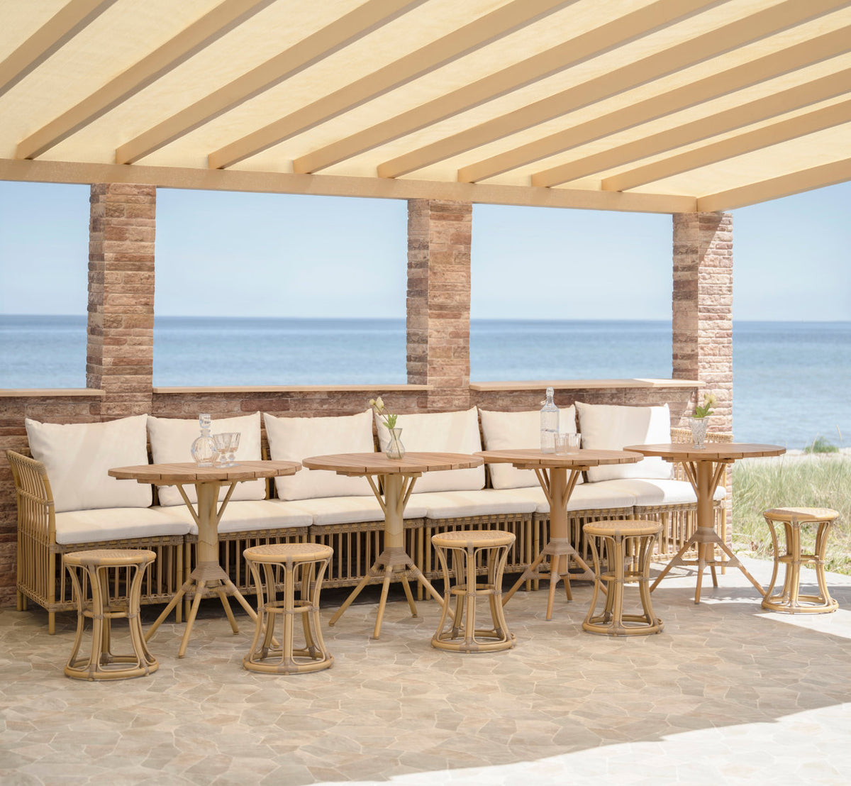 Esta es una imagen de estilo de vida de Bella taburete exterior en natural aluminium artfibre. Banco con taburetes y mesas de café bajo una pérgola.