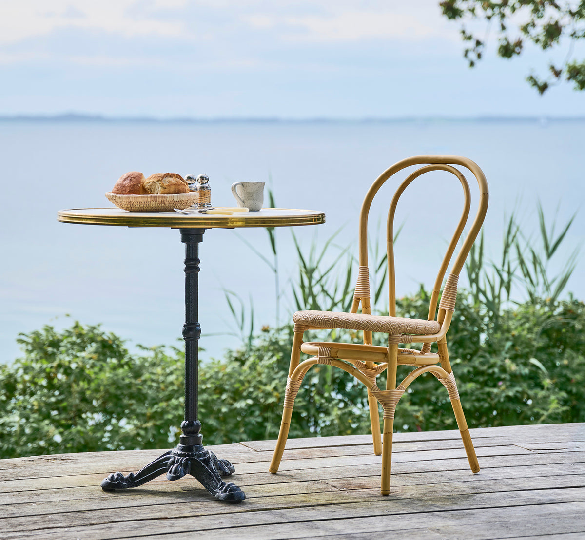 Esta es una imagen de estilo de vida de Lulu silla de comedor exterior en natural aluminium artfibre. En una mesa de bistró frente al mar con pastelería y mar en calma.