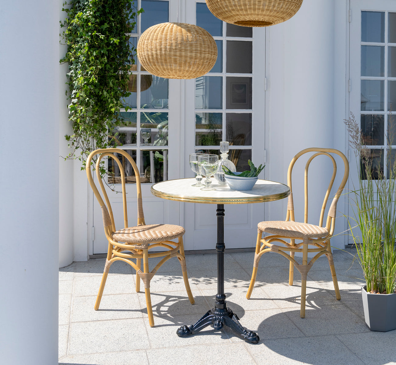 Esta es una imagen de estilo de vida de Lulu silla de comedor exterior en natural aluminium artfibre. En una terraza blanca con una mesa de café y plantas trepadoras.