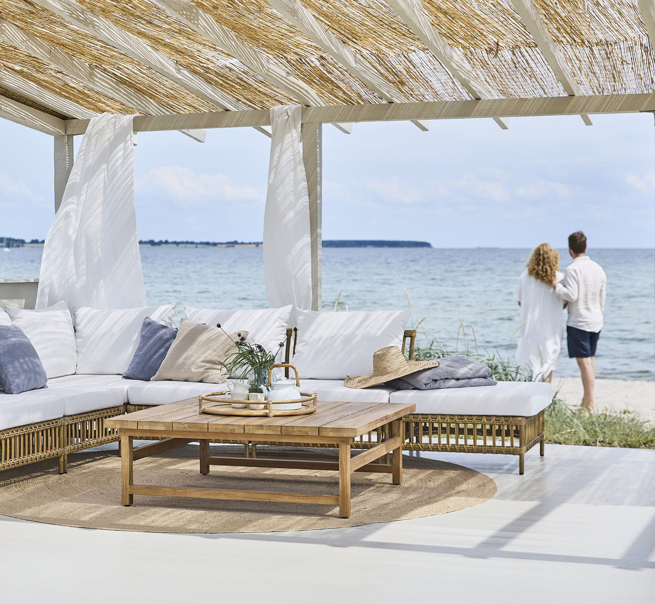 Esta es una imagen de estilo de vida de Maggie Sofá Módulo Puf c. Cristal en antique aluminium artfibre. Pabellón de playa con pareja y vista al mar.