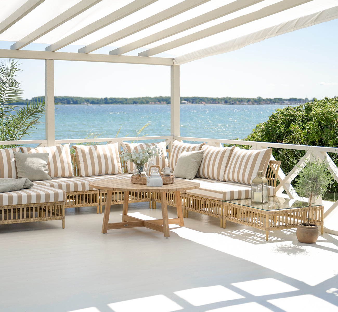 Esta es una imagen de estilo de vida de Maggie Sofá Módulo Puf c. Cristal en natural aluminium artfibre. Terraza cubierta con cojines a rayas y vista al mar.