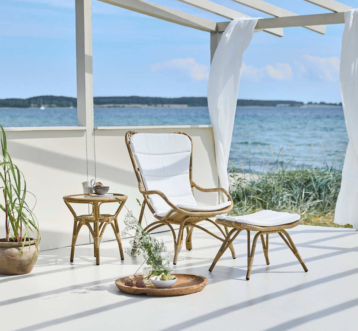 Esta es una imagen de estilo de vida de Monet Exterior Footstool i antique aluminium artfibre. En un pabellón junto al mar con sillón acolchado.