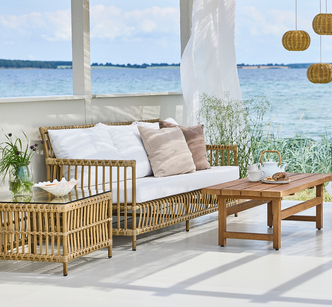 Esta es una imagen de estilo de vida de Caroline sofá exterior en antique Aluminium ArtFibre, en una terraza de madera junto al agua con vegetación.