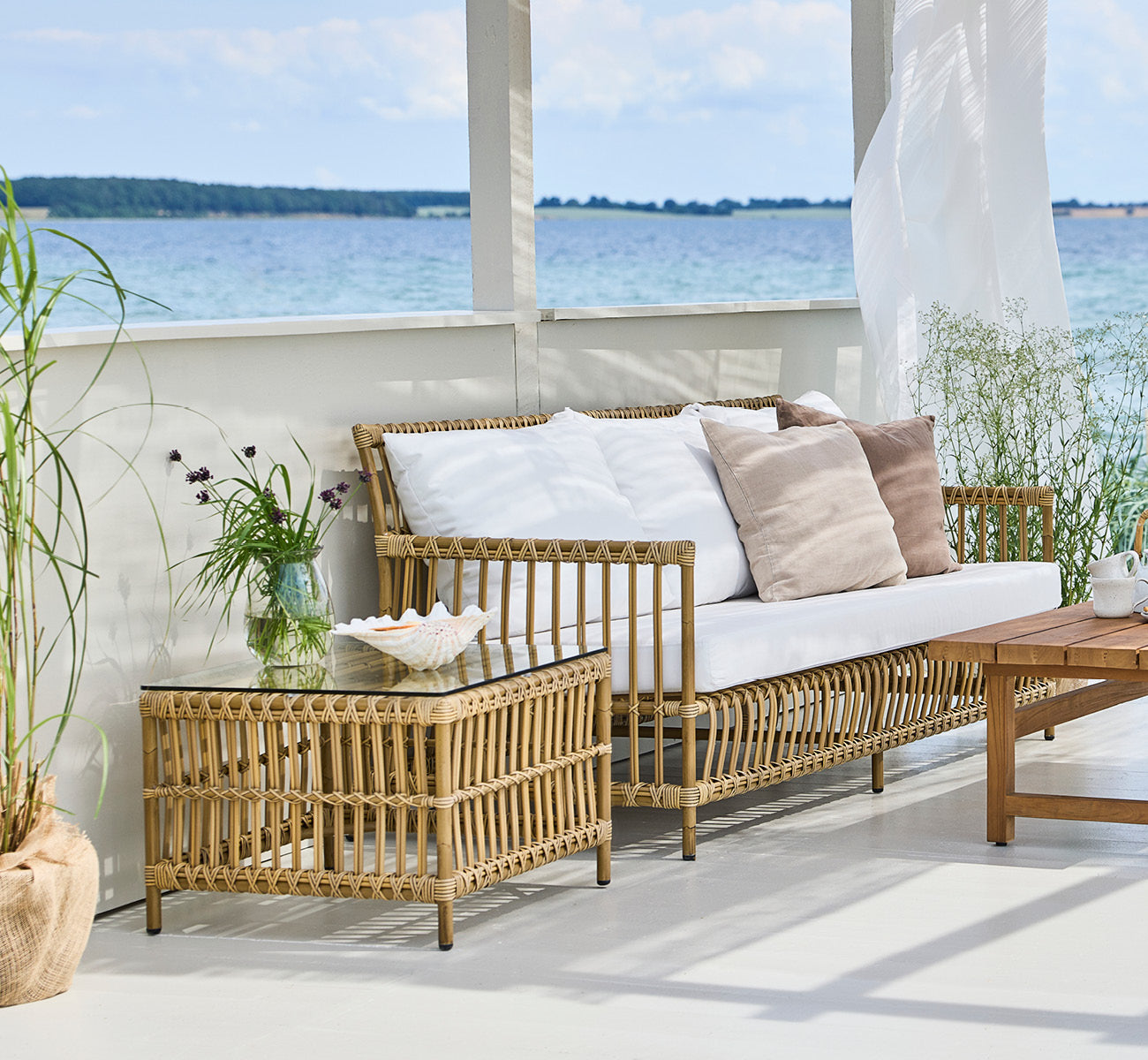 Esta es una imagen de estilo de vida de Caroline mesa exterior en antique Aluminium ArtFibre, en una terraza de madera junto al agua con vegetación.