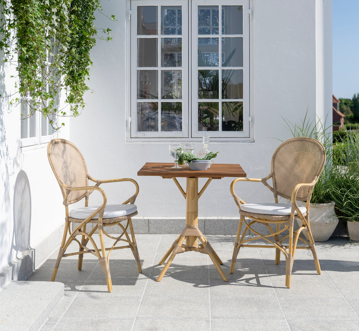 Esta es una imagen de estilo de vida de Nicole Cafe Table i natural aluminium artfibre. En un patio de piedra con sillas Ofelia y vegetación.