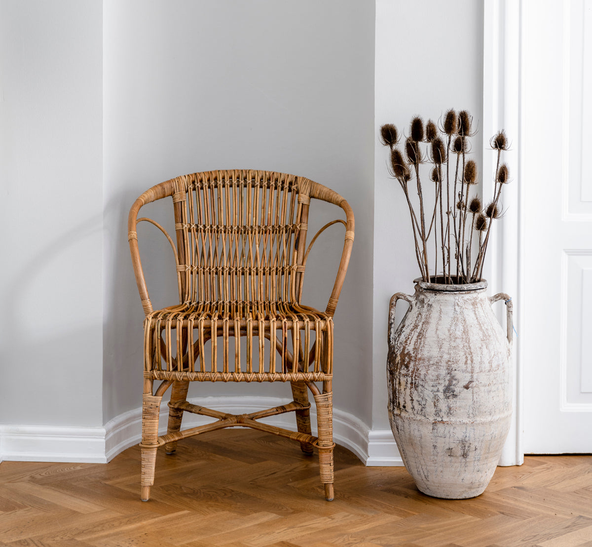 Esta es una imagen de estilo de vida de Robert silla de comedor en polished antique rattan. Colocada junto a una alta urna de terracota sobre un suelo de parqué en espiga.